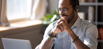 Homme afro-américain du millénaire portant des lunettes et des écouteurs, heureux de regarder un webinaire éducatif sur son ordinateur portable. Un jeune homme d'affaires métis souriant en train de passer un appel vidéo avec ses clients et partenaires.