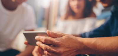 Jóvenes amigos sentados al aire libre y mirando el smartphone. Joven mostrando algo interesante a sus amigos. Enfoque en el teléfono móvil en la mano.